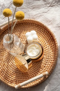 A cosmetic of skincare cream, a decorative candle, and a vase with yellow flowers rest on a woven