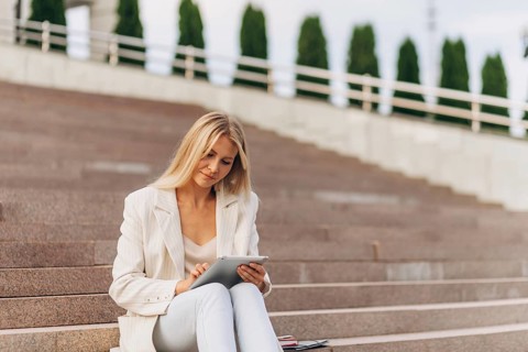 A woman in a white business suit sits on outdoor steps, using a tablet. She appears focused and