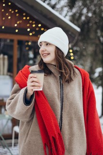 A young woman smiles gently as she holds a cup of coffee outdoors during a snowy winter day. She is