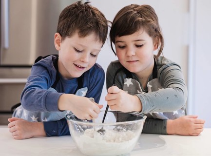 Two young siblings enthusiastically mix ingredients in a bowl, learning to bake in a home kitchen.