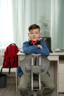 A young boy sits confidently in a chair, wearing a blue shirt with headphones around his neck. A