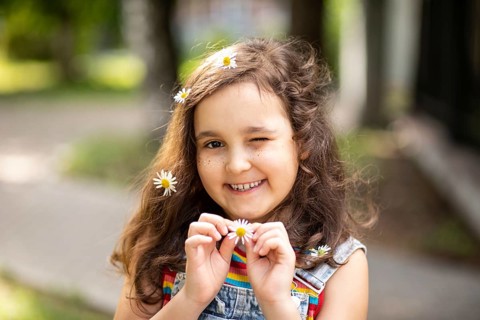 A cheerful little girl smiles warmly, holding a chamomile flower with more in her hair. She stands