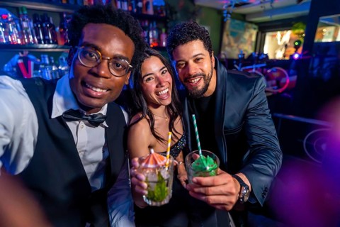 Cheerful multi ethnic friends taking selfie and showing cocktails while enjoying nightlife at