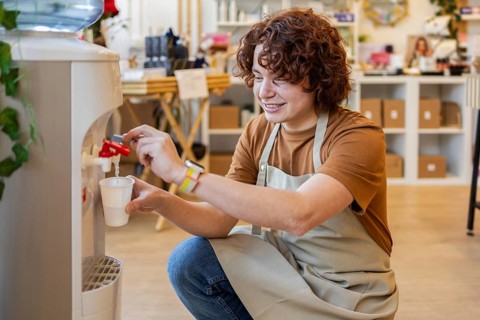 A cheerful shop employee in an apron fills a glass of water from a dispenser while taking a break