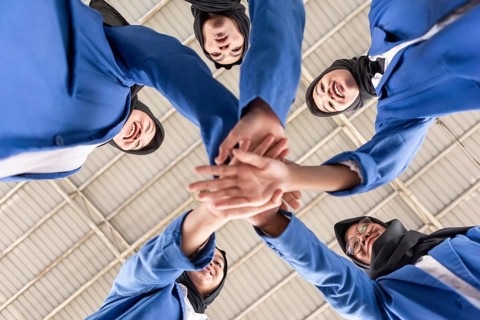 A group of Asian Muslim women in blue uniforms collaboratively join hands, symbolizing unity and