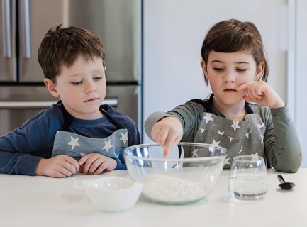 Two siblings wearing star patterned aprons are engaged in baking activities in a modern kitchen.
