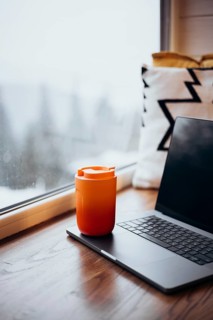 A laptop beside a vibrant orange thermos cup set on a wooden table, illustrating a comfortable
