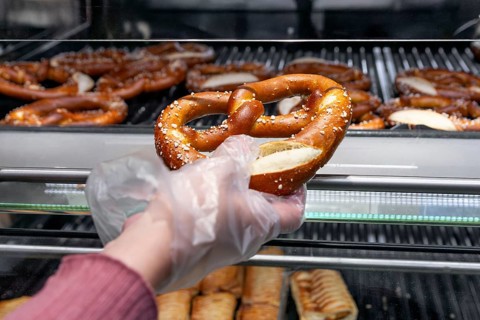 A close up of a freshly baked, salted pretzel being picked up from a bakery shelf with a gloved