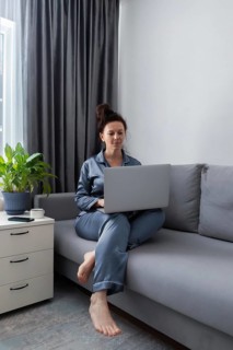 A woman in comfortable pajamas sits on a gray sofa using a laptop, surrounded by a cozy setting