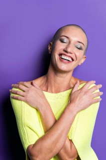 Middle aged shaved head woman in a vibrant yellow sweater, smiling broadly with eyes closed,