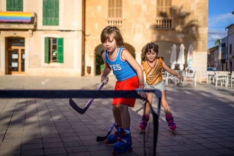 Two children play street hockey on roller blades in a sunlit square. They are focused and