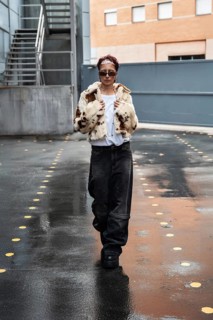 A woman wearing a stylish fur coat and sunglasses walks confidently on a wet catwalk urban street.