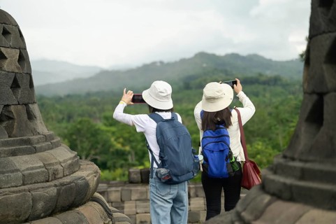 Back view of unrecognizable tourists wearing hats and backpacks, standing at the historic Borobudur