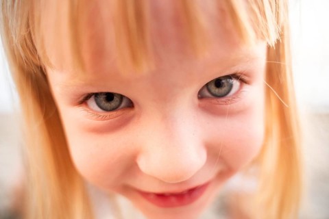 A young girl gazes directly into the camera with bright eyes and a playful smile. This close-up