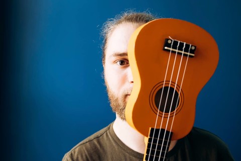 Close-up of a man holding a Mexican ukulele covering half of his face against a vibrant blue