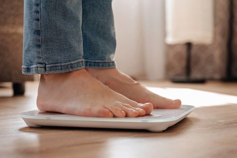 A young woman stands on scales at home, emphasizing a personal weight loss journey. The image