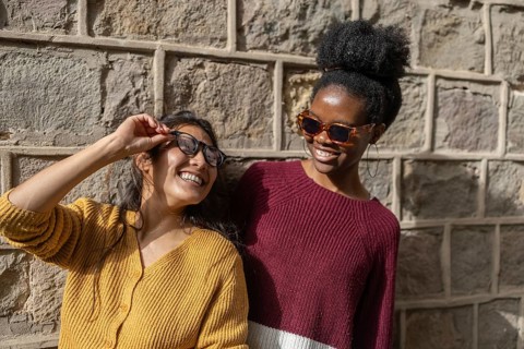 Two friends are laughing and enjoying a sunny day while wearing trendy sunglasses, standing against