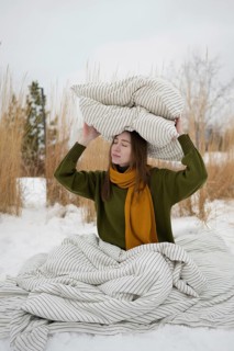 A woman sits on snow, wrapped in a striped blanket, holding a pillow on their head. Wearing a green
