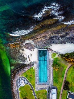 A stunning aerial view captures a seaside pool adjacent to a rocky shore with waves crashing,
