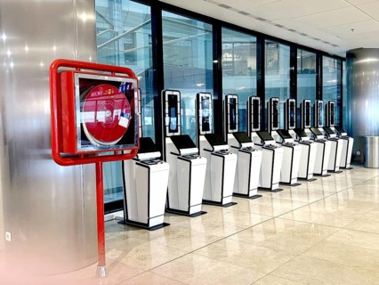 A row of self-service kiosks in a modern airport setting, alongside a fire safety hose reel. The