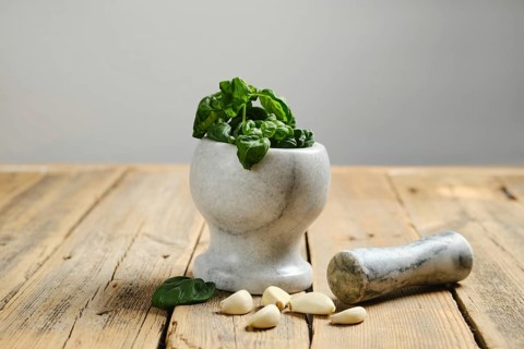 A marble mortar holds fresh green basil ready for pesto preparation. Garlic cloves lie nearby on