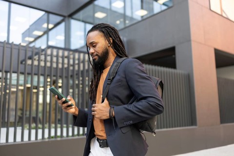 Business man wearing a blazer and backpack, walking outside a modern office building while checking