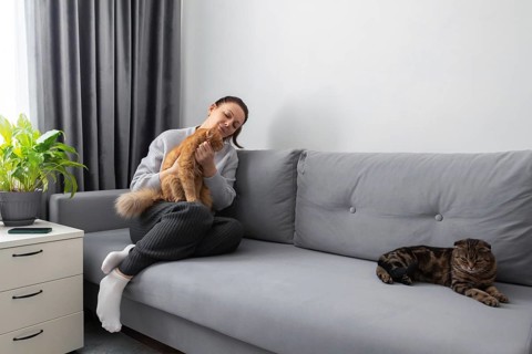 A relaxed woman cuddles her ginger cat on a gray couch, while another cat lounges beside her in a