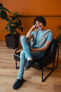 A young man with curly hair sits casually in a modern black chair against an orange backdrop. He's