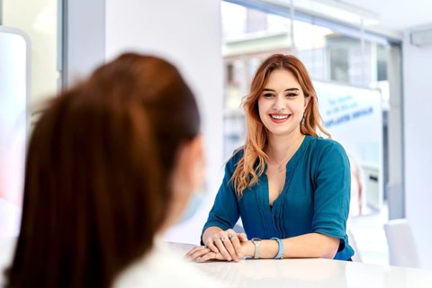 Young woman at a dental clinic reception desk, warmly greeting a visitor. The modern interior and