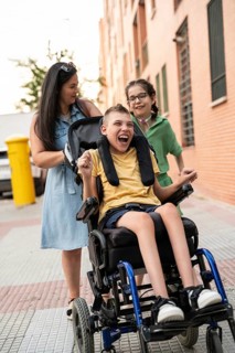 A family enjoys a day out. The mother smiles, pushing her son, who has cerebral palsy, in a