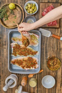 A hand is seen sprinkling a rich herb and spice mixture over fresh fish fillets in a baking tray