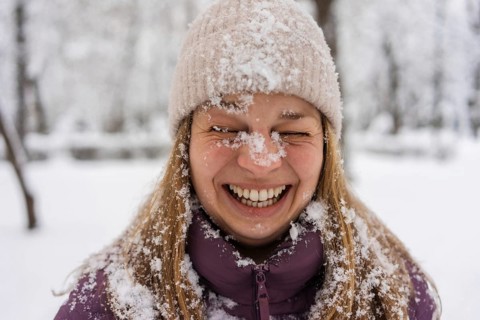 A young woman joyfully laughing, covered in snowflakes. She wears a purple jacket and a knitted
