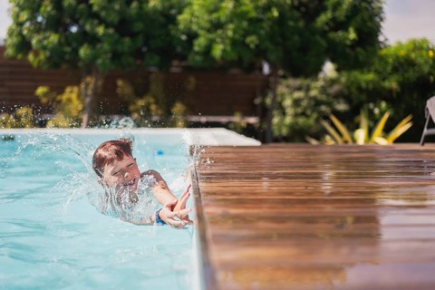 A joyful child plays in a sunlit outdoor pool, splashing water around. Surrounded by lush greenery,