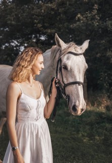 A young woman in a white dress stands beside a white horse, holding its bridle in a sunny meadow.