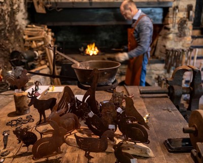 A blacksmith works near a forge in a rustic workshop, with handmade metal animal figures displayed