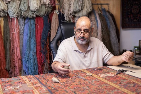 A senior male Iranian master skillfully restores a rare carpet in his well-equipped workshop in