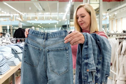 A woman examines a pair of jeans in a contemporary store filled with clothing. She inspects the