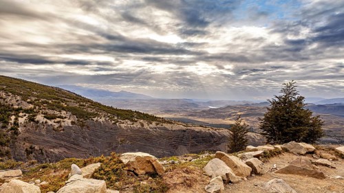 Rocks and trees with sweeping views of mountains and the vast landscape below under a cloudy sky,