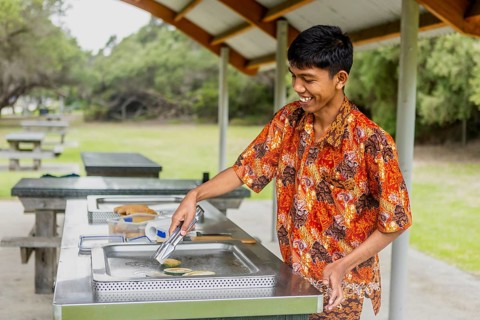 An Indonesian man wearing vibrant batik clothing enjoys cooking at an outdoor barbecue in Australia