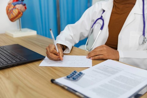 A doctor is seen writing notes on paper in a medical office. The scene includes a laptop,