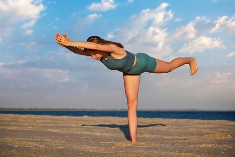 A woman gracefully performs the Virabhadrasana Warrior 3 pose on a sandy beach as the sun sets in
