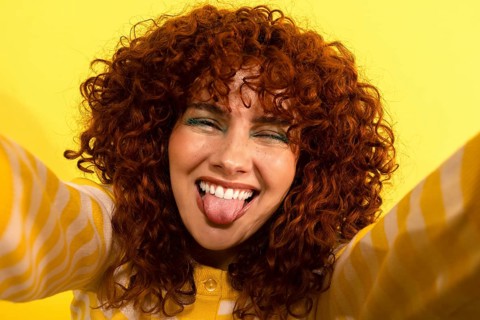 A cheerful woman with vibrant curly red hair playfully sticks out her tongue while making a silly