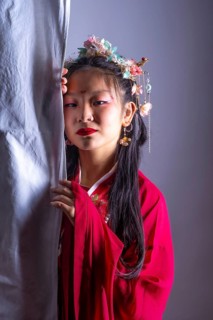 A young girl in a red dress with a flower headband stands behind a curtain. She has red lipstick on