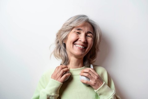 Asian senior woman with grey hair smiling joyfully, wearing green sweater and headphones,