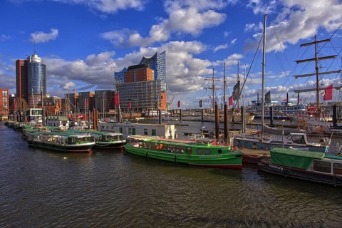 View of boats and ships in harbor surrounded by city skyline under a partly cloudy blue sky,