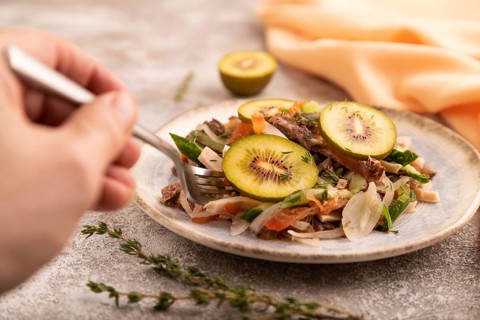 Meat salad with beef kiwi and vegetables with hand on brown concrete background and orange linen
