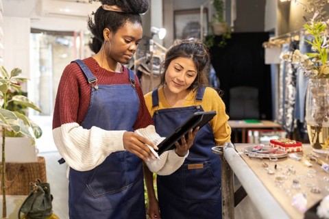 Two women in a fashion retail store, wearing blue aprons, collaborate using a digital tablet. They
