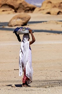Woman in traditional dress carrying objects on her head, surrounded by a desert-like landscape,
