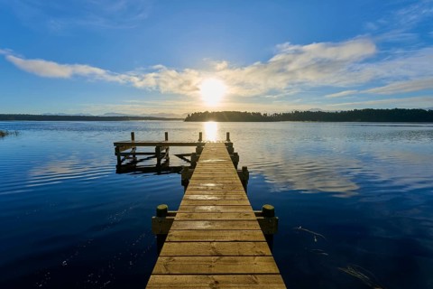 A wooden jetty leads into the calm waters of a lake, the sun is low in the sky, Lake Mahinapua,