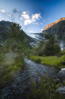A tranquil stream winds through lush greenery, fed by a glacier nestled in the Norwegian mountains